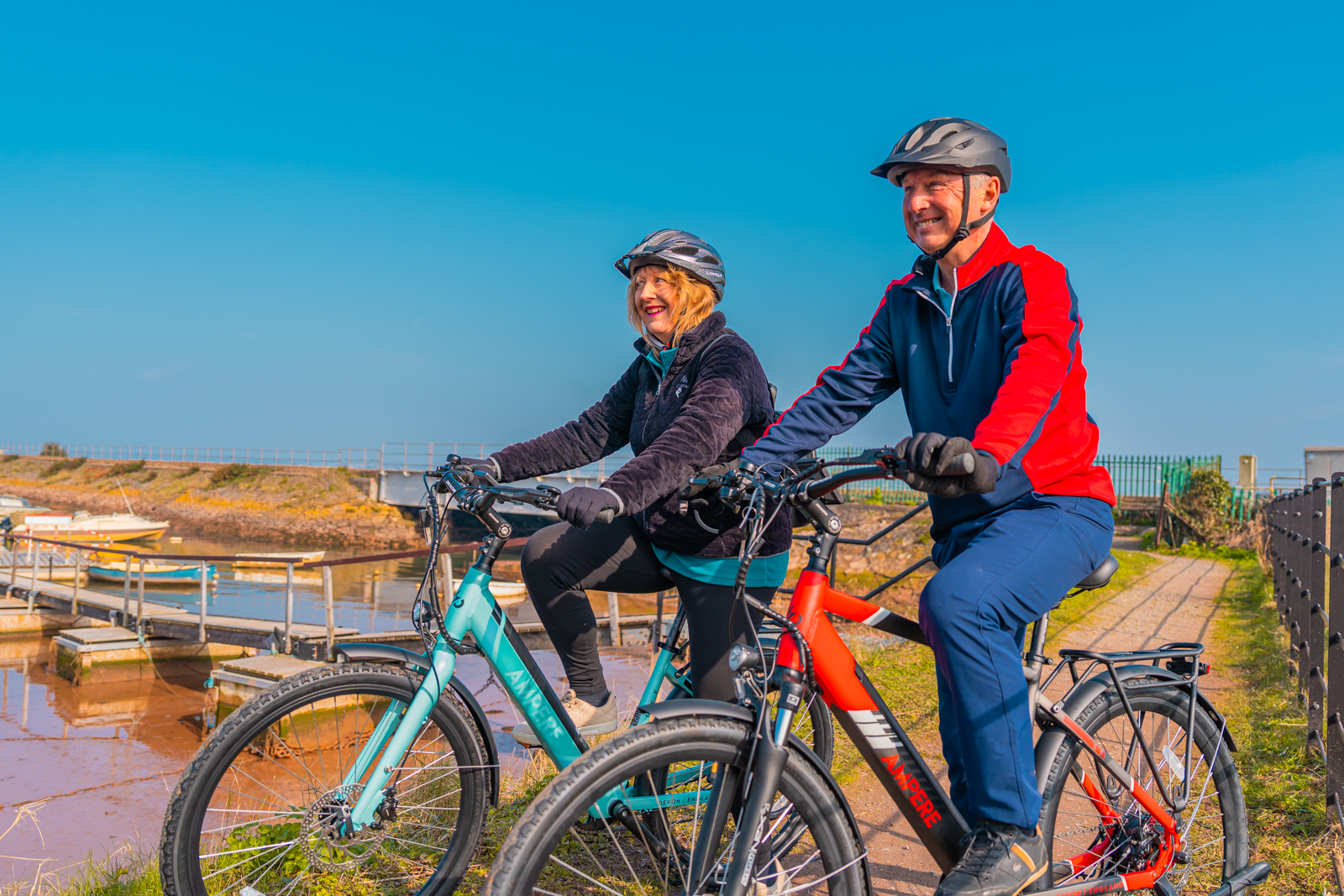 Couple riding Ampere electric bikes along a waterfront path, available through all UK cycle schemes